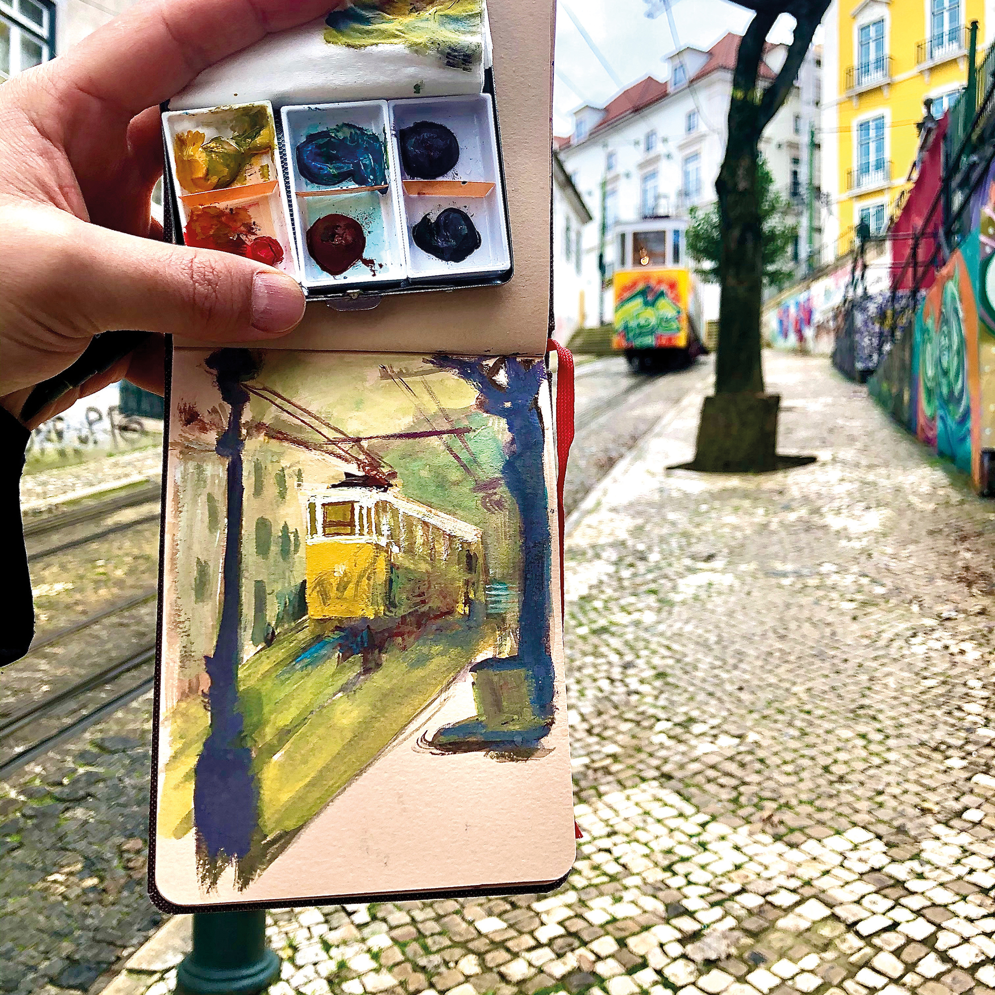 Uma mão segura um potinho com tintas e uma página de caderninho; ao fundo, se vê a vista da rua que está desenhada na página do caderninho.